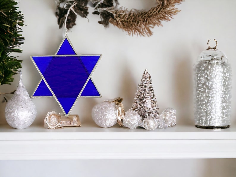 May include: A blue stained glass Star of David ornament on a white shelf with silver and white Christmas ornaments and a silver and white Christmas tree.