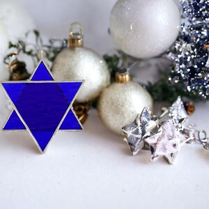 May include: A blue glass Star of David ornament with silver trim, surrounded by white and silver Christmas ornaments.