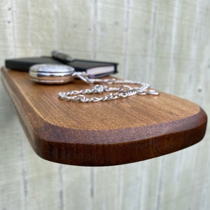 K&ouml;nnte beinhalten: Ein Holzregal mit abgerundeter Kante, in einem dunklen Braunton gebeizt. Ein schwarzes Notizbuch, eine silberne Taschenuhr und eine silberne Kette liegen auf dem Regal.