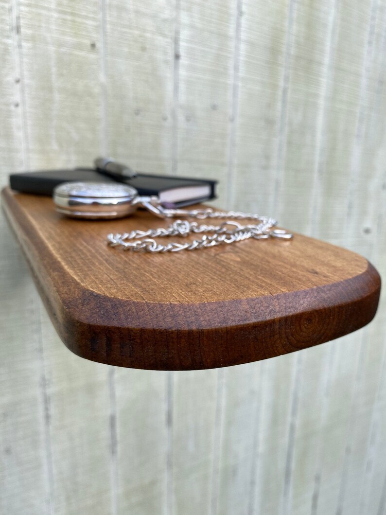 K&ouml;nnte beinhalten: Ein Holzregal mit abgerundeter Kante, in einem dunklen Braunton gebeizt. Ein schwarzes Notizbuch, eine silberne Taschenuhr und eine silberne Kette liegen auf dem Regal.