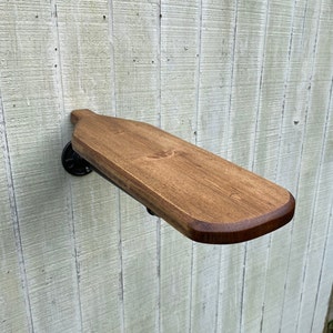 K&ouml;nnte beinhalten: Ein Holzregal mit abgerundeter Kante, das mit einer schwarzen Metallrohrhalterung an einer Wand befestigt ist. Das Regal ist in einem dunklen Braunton gebeizt.