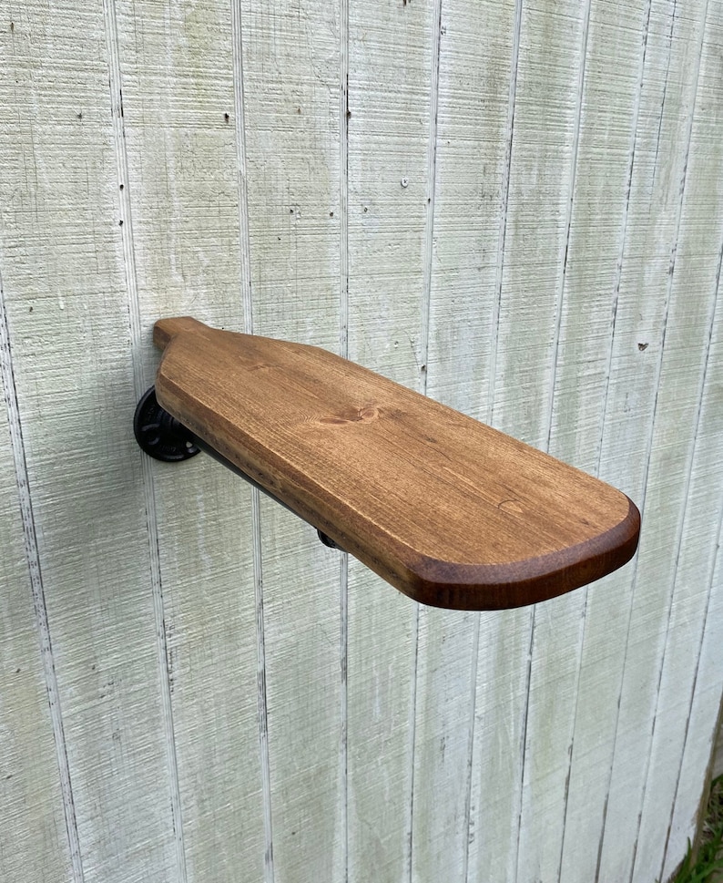 K&ouml;nnte beinhalten: Ein Holzregal mit abgerundeter Kante, das mit einer schwarzen Metallrohrhalterung an einer Wand befestigt ist. Das Regal ist in einem dunklen Braunton gebeizt.