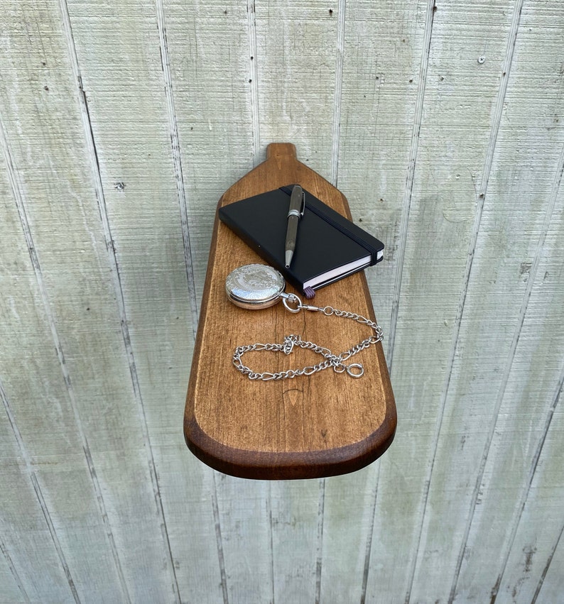 K&ouml;nnte beinhalten: Ein Holzregal in Paddelform mit einem schwarzen Notizbuch, einem silbernen Stift und einer silbernen Taschenuhr mit Kette.