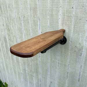 K&ouml;nnte beinhalten: Ein Holzregal mit abgerundeter Kante, das mit schwarzen Metallrohrhalterungen an einer Wand befestigt ist. Das Regal ist in einem dunklen Braunton gebeizt.