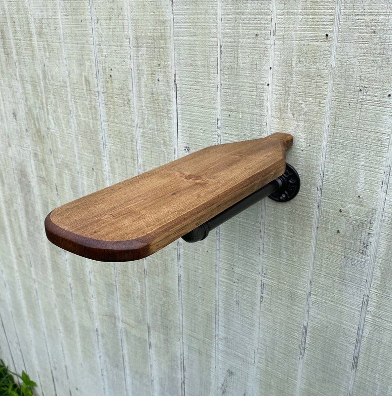 K&ouml;nnte beinhalten: Ein Holzregal mit abgerundeter Kante, das mit schwarzen Metallrohrhalterungen an einer Wand befestigt ist. Das Regal ist in einem dunklen Braunton gebeizt.