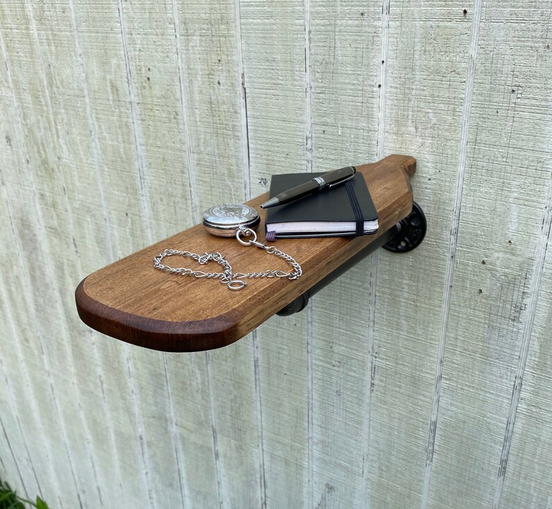 K&ouml;nnte beinhalten: Ein Holzregal mit schwarzen Metallrohrst&uuml;tzen. Das Regal ist an einer wei&szlig;en Holzwand montiert. Eine Taschenuhr, ein Stift und ein Notizbuch liegen auf dem Regal.