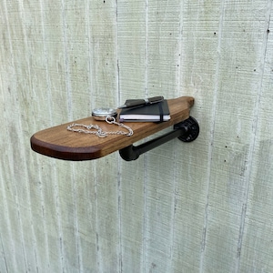 K&ouml;nnte beinhalten: Ein Holzregal mit einer schwarzen Metallrohrst&uuml;tze. Das Regal ist an einer wei&szlig;en Wand montiert und hat ein Notizbuch, einen Stift und eine Taschenuhr darauf.