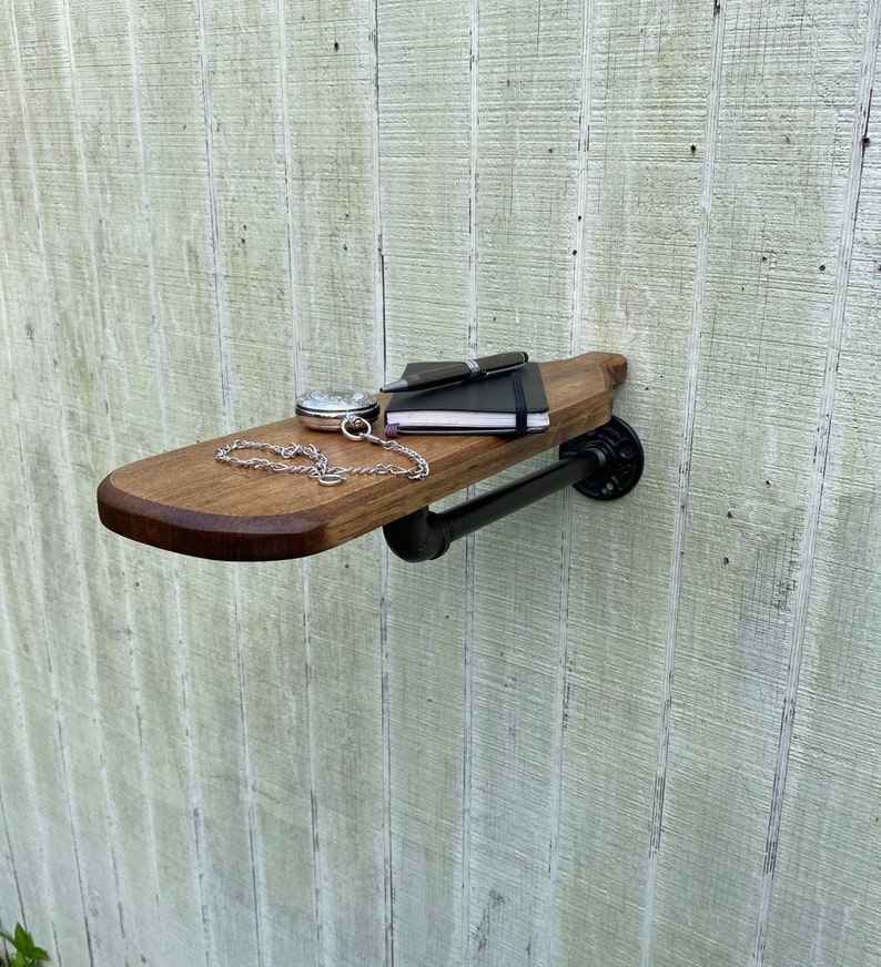 K&ouml;nnte beinhalten: Ein Holzregal mit einer schwarzen Metallrohrst&uuml;tze. Das Regal ist an einer wei&szlig;en Wand montiert und hat ein Notizbuch, einen Stift und eine Taschenuhr darauf.