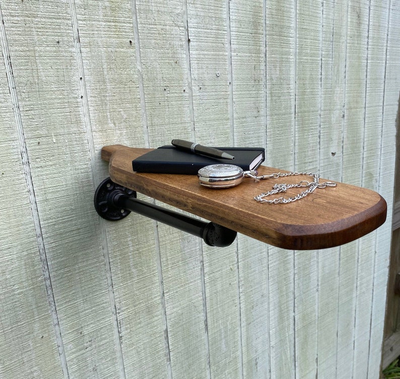 K&ouml;nnte beinhalten: Ein Holzregal mit schwarzen Metallrohrhalterungen. Das Regal ist an einer wei&szlig;en Wand montiert und hat ein Notizbuch, einen Stift und eine Taschenuhr darauf.