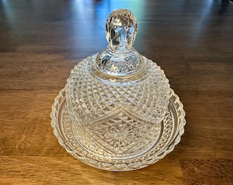 Può includere: Una burriera in vetro tagliato trasparente con coperchio a cupola e base rotonda. Il coperchio presenta un pomello sfaccettato e un motivo a taglio diamante. La base ha un bordo smerlato e un disegno a stella al centro. Il piatto è progettato per contenere burro.