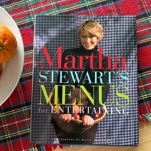 May include: A cookbook titled "Martha Stewart's Menus for Entertaining" is displayed on a plaid tablecloth. The cover features a photo of Martha Stewart holding a wooden bowl of fruit. The book's title is in large, colorful letters.