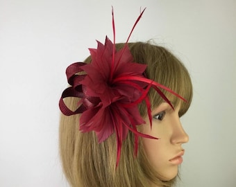 May include: A burgundy floral hair accessory. The headpiece features a large, layered flower with delicate petals and long, thin, red accents. A looped ribbon adds texture. Suitable for special occasions, it's a stylish addition.