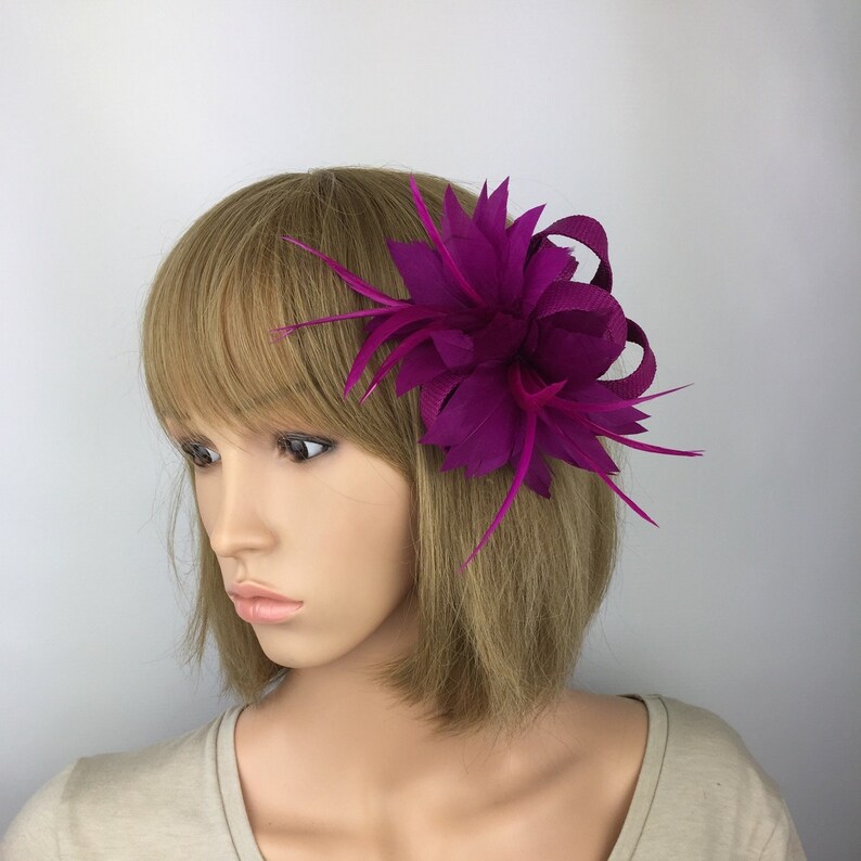 May include: A magenta feather flower hair clip. The flower is made of feathers and has a ruffled texture. The clip is attached to the back of the flower.