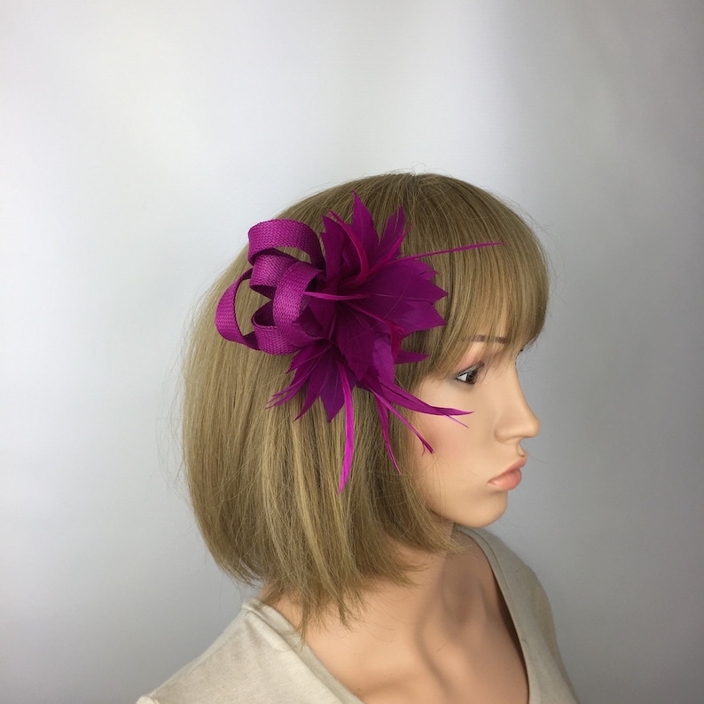 May include: A magenta flower hair clip with a satin ribbon and feathers. The clip is worn on a mannequin with short brown hair.