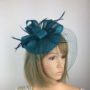 May include: Teal blue fascinator with a large bow and a veil. The fascinator is made of a soft, textured fabric and has a small, round base.