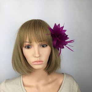 May include: A purple flower hair clip with a feather accent. The clip is worn on a mannequin with short brown hair.
