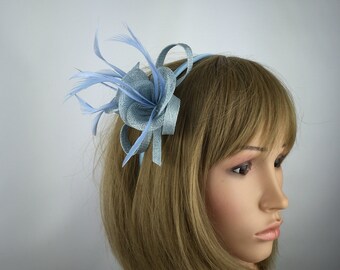 May include: A pale blue headpiece with a floral and feather design. The headpiece features a central flower shape with layered petals and delicate feathers. The headpiece is made of fabric and is worn on a mannequin.