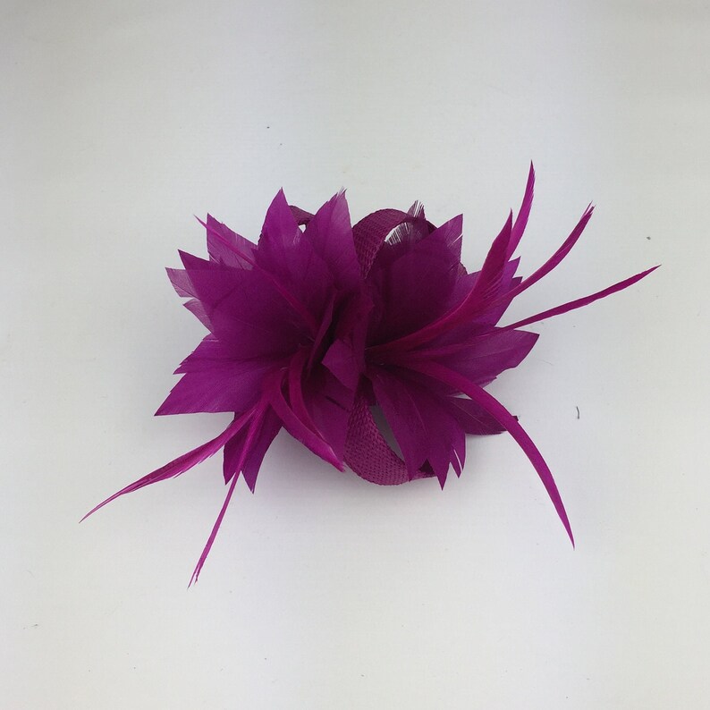May include: A magenta feather flower hair clip. The flower is made of multiple layers of feathers and has a small ribbon in the centre.