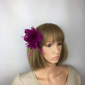 May include: A magenta flower hair clip with a single feather accent. The clip is worn on a mannequin with short brown hair.