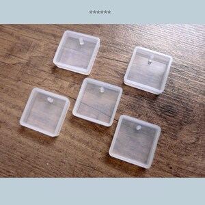 Könnte beinhalten: Fünf klare, quadratische Silikonformen mit einer kleinen Schlaufe oben zum Basteln. Die Formen sind auf einer Holzoberfläche angeordnet. Die Formen sind durchscheinend und scheinen leer zu sein.