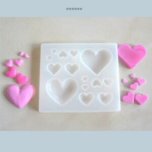 Puede incluir: Un molde de silicona blanco con m&uacute;ltiples cavidades en forma de coraz&oacute;n de diferentes tama&ntilde;os. El molde est&aacute; rodeado de piezas de resina en forma de coraz&oacute;n rosa.