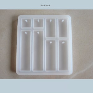 Könnte beinhalten: Eine transparente Silikonform mit acht rechteckigen Vertiefungen zum Herstellen von Schmuckanhängern. Jede Vertiefung hat ein kleines Loch oben zum Anbringen eines Sprungrings oder einer Öse.