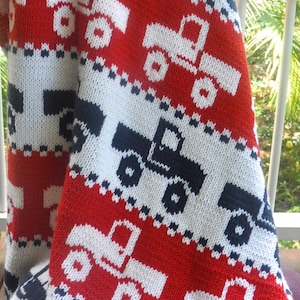 May include: A knitted baby blanket with a red, white, and blue pattern featuring a repeated design of trucks. The blanket is made of soft yarn and is perfect for a baby boy or girl.