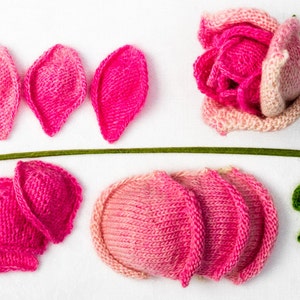 May include: A collection of knitted flower components, including petals in varying shades of pink, a green stem, and leaves. The assembled flower is a vibrant pink and light pink rose.