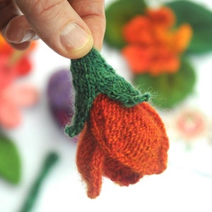 May include: Close-up of a handmade, knitted orange flower with a green stem. The flower is held in a person's hand, with other colorful knitted flowers in the background. The craft is likely for decoration.