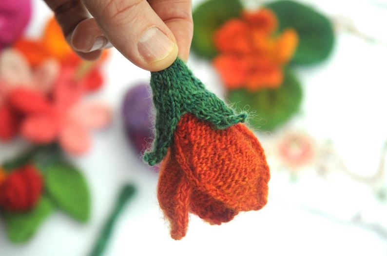 May include: Close-up of a handmade, knitted orange flower with a green stem. The flower is held in a person's hand, with other colorful knitted flowers in the background. The craft is likely for decoration.