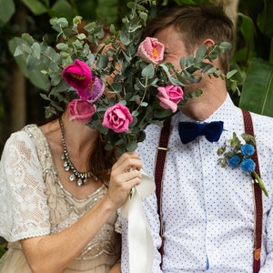 May include: A bouquet of pink and fuchsia crocheted flowers with green foliage, held in front of a couple. The man wears a white shirt with blue polka dots, a navy bow tie, and burgundy suspenders. The woman wears a cream-colored dress with lace sleeves.