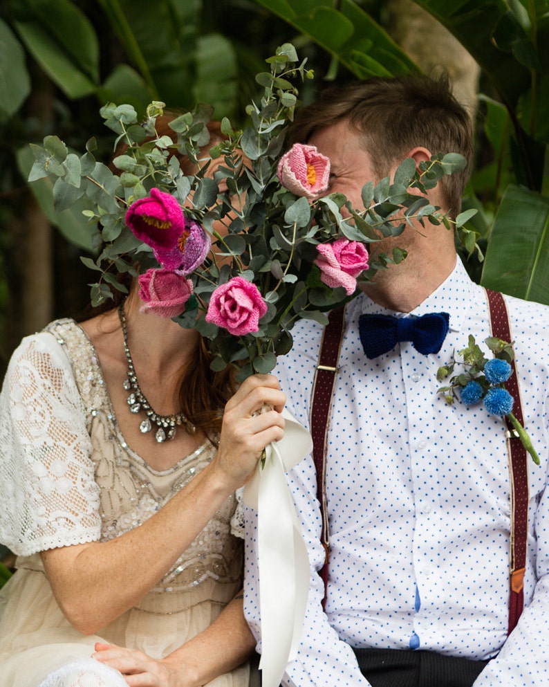 May include: A bouquet of pink and fuchsia crocheted flowers with green foliage, held in front of a couple. The man wears a white shirt with blue polka dots, a navy bow tie, and burgundy suspenders. The woman wears a cream-colored dress with lace sleeves.
