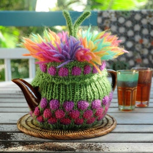 May include: A green knitted teapot cozy with pink polka dots and a rainbow-colored fluffy top. The cozy is on a brown wicker coaster.