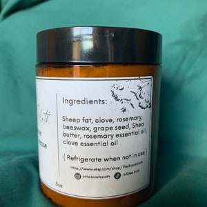 Pu&ograve; includere: Un barattolo di vetro marrone con un coperchio nero, etichettato "Ingredienti: Grasso di pecora, chiodi di garofano, rosmarino, cera d'api, semi d'uva, burro di karit&egrave;, olio essenziale di rosmarino, olio essenziale di chiodi di garofano". Il barattolo include anche il testo "Refrigerare quando non in uso" e un indirizzo del sito web. Il barattolo &egrave; etichettato "8oz".