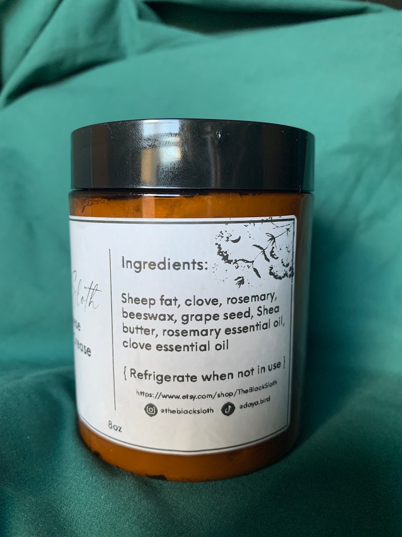 Pu&ograve; includere: Un barattolo di vetro marrone con un coperchio nero, etichettato "Ingredienti: Grasso di pecora, chiodi di garofano, rosmarino, cera d'api, semi d'uva, burro di karit&egrave;, olio essenziale di rosmarino, olio essenziale di chiodi di garofano". Il barattolo include anche il testo "Refrigerare quando non in uso" e un indirizzo del sito web. Il barattolo &egrave; etichettato "8oz".