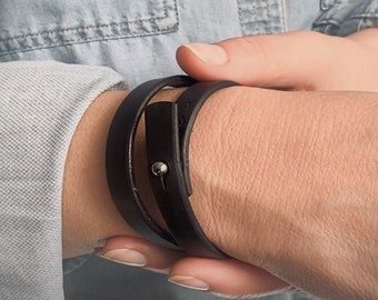 May include: A black leather wrap bracelet with a silver metal accent. The bracelet is worn on a person's wrist.