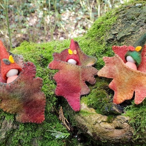 May include: Three handmade felt leaf ornaments with small wooden doll figures. Each leaf is a different colour, ranging from red to orange, with a stitched border. The dolls wear tiny hats in various colours, including red and green.