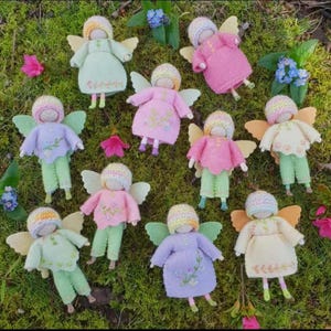 Op de afbeelding: Een verzameling handgemaakte feeënpoppen met garen haar, vleugels en outfits in verschillende pastelkleuren. De poppen zijn gerangschikt op groene mos met verspreide bloemen, wat een grillige scène creëert.