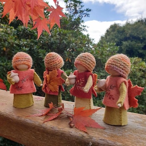May include: Four handmade felt dolls in autumnal hues, each holding a small acorn. They wear knitted hats, felt vests with leaf and branch designs, and felt skirts. Red and orange autumn leaves are scattered around them.