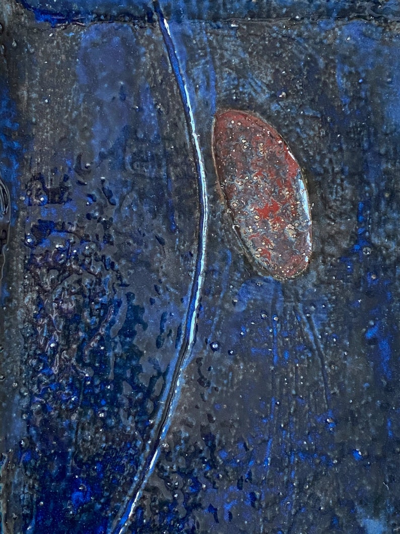 Pu&ograve; includere: Primo piano di una piastrella di ceramica blu con una forma ovale di colore rosso ruggine al centro. La piastrella ha una superficie testurizzata e una linea bianca che attraversa in diagonale.