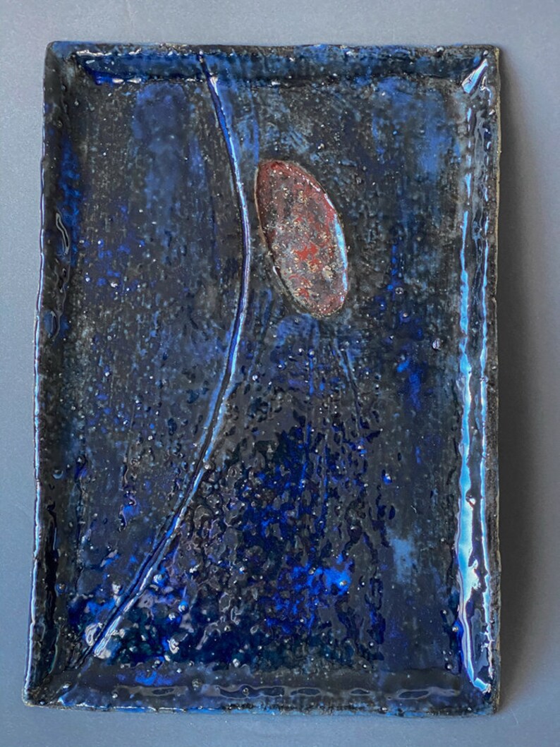Pu&ograve; includere: Un vassoio rettangolare in ceramica con una glassa blu scuro e una singola forma ovale rosso-marrone al centro. Il vassoio ha una superficie testurizzata e un bordo leggermente rialzato.