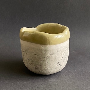 May include: A small, ceramic cup with a textured, white exterior and a light green rim. The cup has a slightly irregular shape.