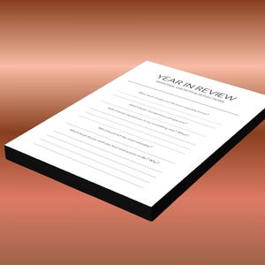 May include: A white Year in Review journal with black edges, featuring prompts for personal growth and reflections. The journal is set against a gradient brown background. The text on the journal includes questions about books and life.