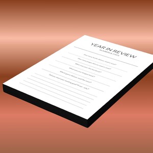 May include: A white Year in Review journal with a black border, lying on a gradient brown background. The journal has lines for recording book-related statistics, such as the number of books read and the longest book.