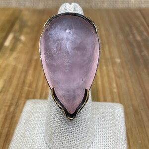 May include: A pink teardrop-shaped ring with a gold setting. The ring is on a white fabric display stand.