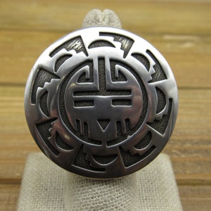 May include: A silver ring with a detailed, circular design featuring a geometric pattern. The ring is set on a white fabric surface.