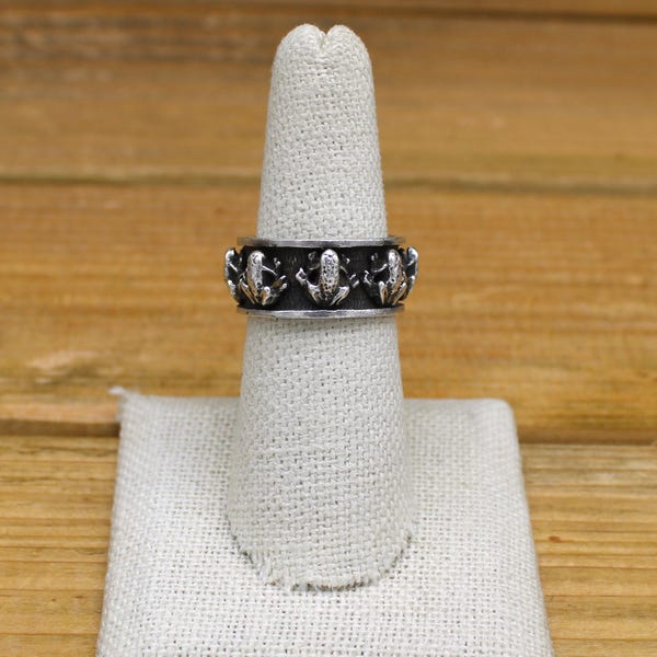 May include: A silver ring with a band of frogs around the circumference. The frogs are facing outwards and have a textured finish.