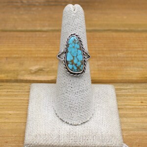 May include: A silver ring with a turquoise stone. The turquoise stone is oval-shaped with a light blue color and brown matrix. The ring has a twisted rope design around the stone and a simple band.