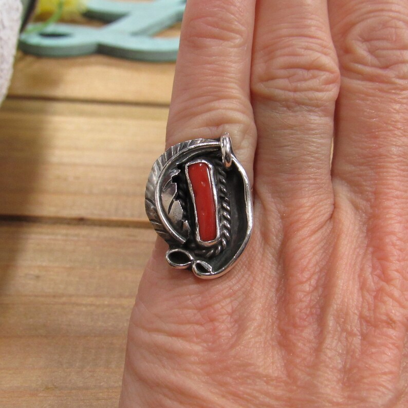 May include: A silver ring with a red coral stone set in a stylized design. The ring is worn on a finger.
