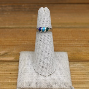May include: A silver ring with four inlaid stones. The stones are blue, turquoise, green, and silver. The ring is displayed on a white ring stand.